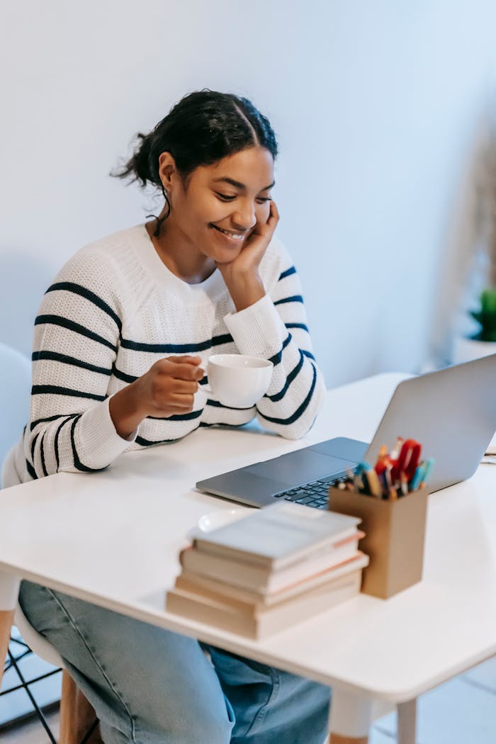 Positive young African American female freelancer in casual clothes talking on smartphone and drinking coffee while working remotely on laptop at home